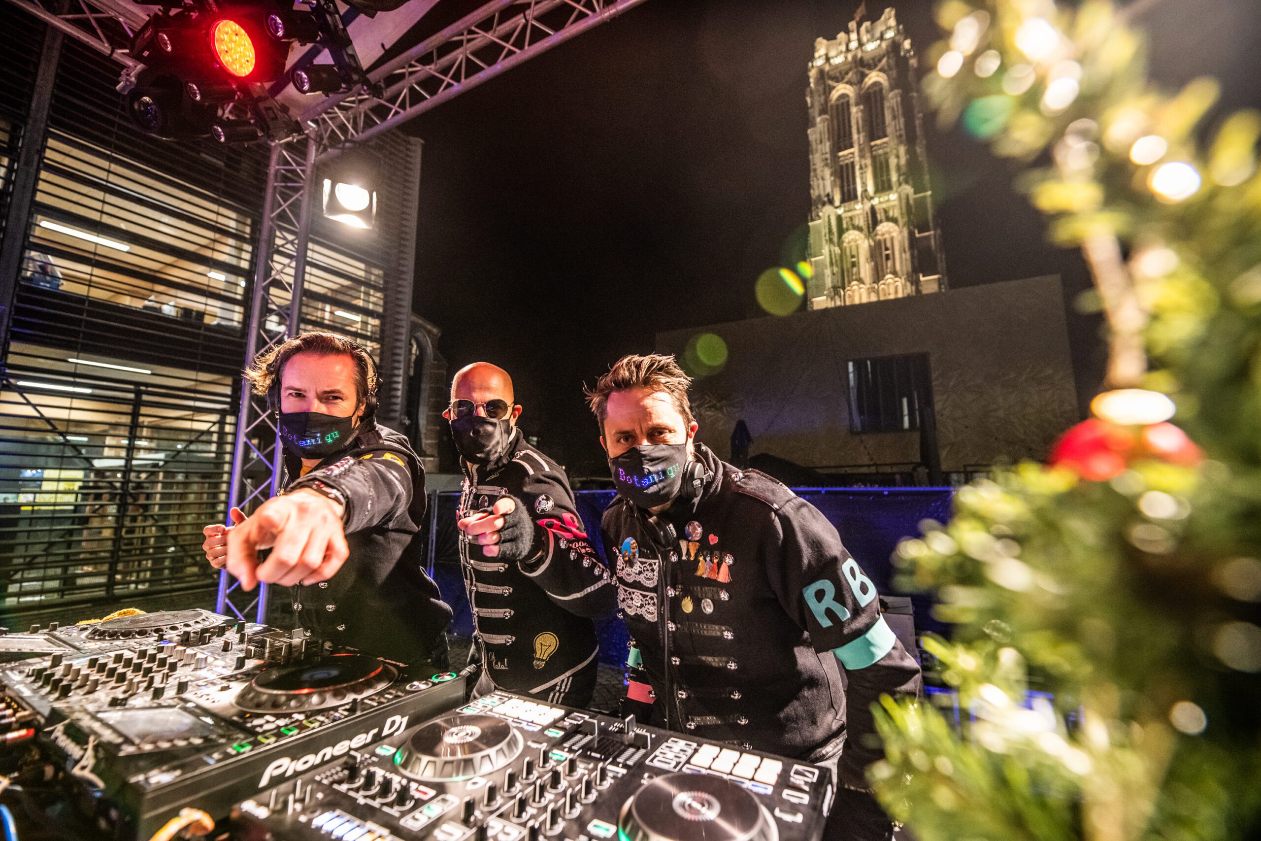 Drie DJ's met mondmasker staan op een podium achter hun DJ-tafel met de Sint-Romboutstoren op de achtergrond.